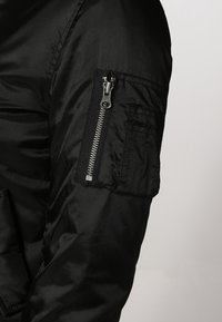 Blouson aviateur noir avec une texture lisse et brillante. Comprend une poche zippée sur le bras et une fermeture éclair proéminente à l'avant.