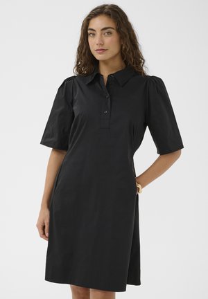 Žena s kučeravými vlasmi v čiernych šatách do kolien s nafúknutými krátkymi rukávmi a zapínaným golierom, stojaca pred jednoduchým pozadím.