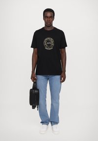 T-shirt en coton noir avec un logo circulaire doré, associé à un jean bleu clair et des baskets blanches, tenant un sac zippé noir.