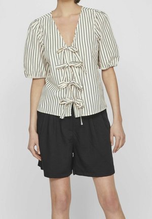 Femme portant un chemisier crème à rayures verticales noires avec quatre nœuds noués sur le devant, associé à un short noir ample.