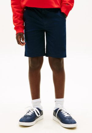 Personne portant un short bleu marine, des chaussettes blanches, des baskets bleu marine avec des lacets blancs et un haut rouge à manches longues, les mains dans les poches.