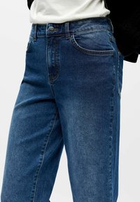 Tmavě modré džínové kalhoty s hladkou texturou, klasickým designem s pěti kapsami, kovovým knoflíkem a prošívanými detaily.