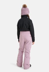 Zwarte longsleeve shirt, lichtroze sneeuwbroek en een roze gebreide muts met een pompon. De sneeuwbroek heeft een logo op de achterkant.