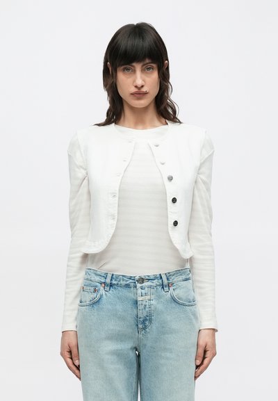 Veste courte en denim blanc avec cinq boutons noirs, portée par-dessus une chemise à manches longues à rayures claires et associée à un jean bleu clair.