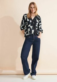 Blusa a maniche lunghe nera e blu chiaro con motivo, con scollatura a V, abbinata a jeans larghi blu scuro e sneaker bianche.