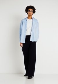 Himmelblau gestreiftes Button-down-Hemd über einem weißen T-Shirt, kombiniert mit schwarzen weiten Hosen, getragen mit Sonnenbrille und schwarzen Schuhen.