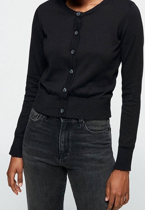 Femme portant un cardigan noir ajusté boutonné avec des bords festonnés et un jean gris foncé taille haute sur un fond uni.