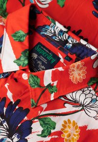 Camisa corta de mangas con estampado floral rojo que presenta patrones en azul, verde y blanco. Tiene cuello con solapa, cierre de botones y una etiqueta de marca en el interior.