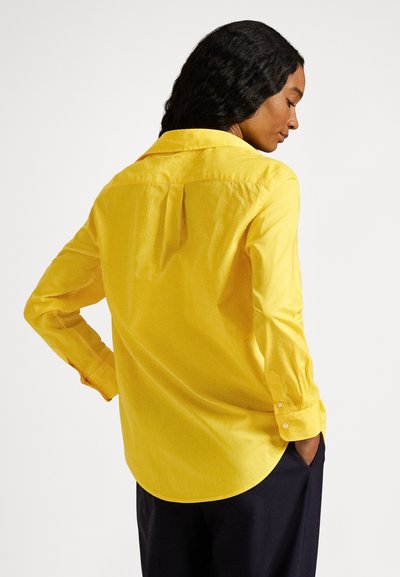 Moteris su tamsiais garbanotais plaukais, apsirengusi ryškiai geltonu ilgomis rankovėmis marškinėliu ir tamsiomis kelnėmis, stovinti šonu, viena ranka remiasi į klubą.