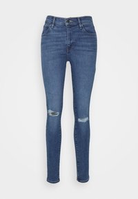 Jeans ajustados de mezclilla azul oscuro con rodillas desgastadas, diseño de cinco bolsillos, pasadores para cinturón y cierre de cremallera.