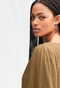 Braune Bluse mit strukturierter Oberfläche, Puffärmeln und gepunktetem Muster, kombiniert mit silbernen Creolen. Haare zu langen Zöpfen geflochten.