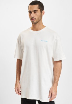 Mann mit kurzem Bart und Haar, der ein schlichtes weißes oversized T-Shirt mit kleinem blauem „JUST RHYSE“-Text auf der Brust vor weißem Hintergrund trägt.