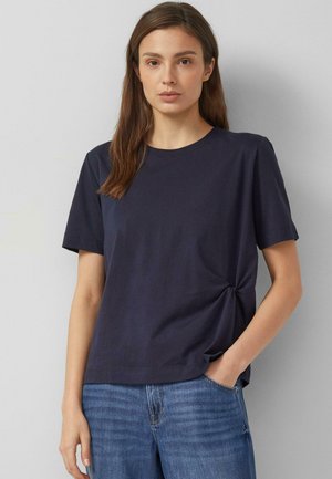 Młoda kobieta o prostych brązowych włosach ubrana w granatowy t-shirt z krótkim rękawem z wiązaniem z boku oraz niebieskie dżinsy, stoi na tle jednolitego tła.