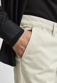 Svetle béžové chino kalhoty s rovnym střihem, které mají šikmé přední kapsy a decentní švy. Nošeny s černou košilí s dlouhým rukávem.