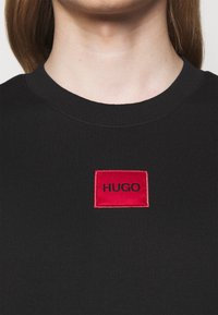 Maglione nero con colletto a costine, caratterizzato da una toppa rettangolare rossa ricamata con la parola "HUGO" in nero. Tessuto liscio.