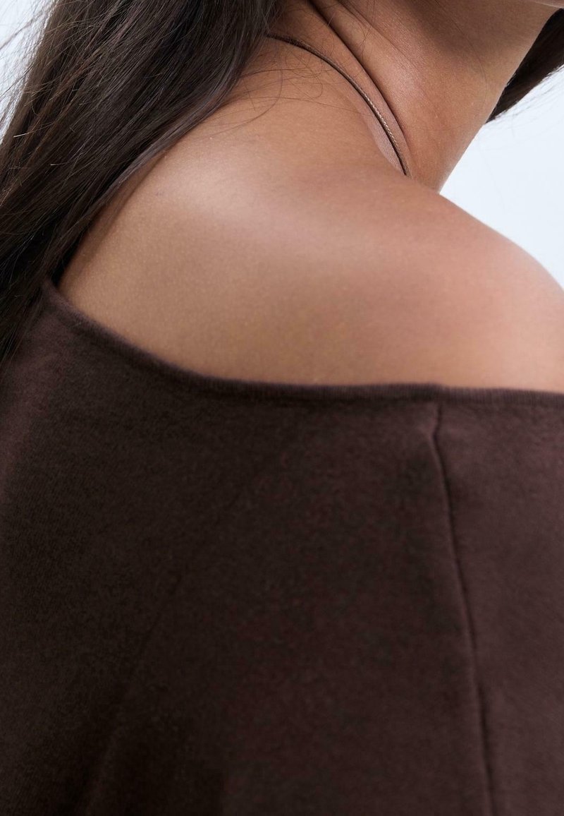 Gros plan d’une épaule nue d’une personne aux cheveux foncés, portant un haut sombre à épaules dénudées sur un fond clair uni.