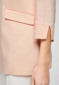 Persikanvärinen bleiseri, jossa on ryhdikäs muotoilu, diagonaalinen tasku vasemmalla puolella ja halkiohiharesorin yksityiskohta.