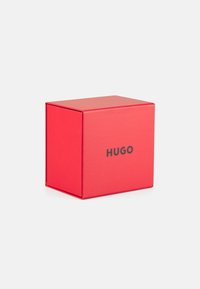 Caja de cartón roja con un acabado mate, que presenta un logo impreso en negro "HUGO" en la parte frontal. Forma cuadrada, sin patrones o acentos adicionales.