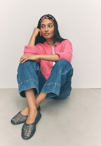 Gestrickte rosa Strickjacke, weißes Top, weite blaue Jeans aus Denim und schwarze gewebte Ballerinas. Modell sitzt vor hellem Hintergrund.