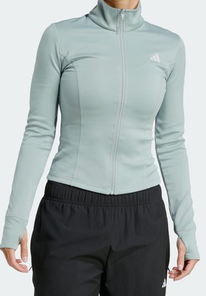 Veste de sport ajustée bleu clair avec fermeture éclair et trous pour les pouces, associée à un pantalon noir ample à taille élastique, portée par une personne visible du cou aux hanches.