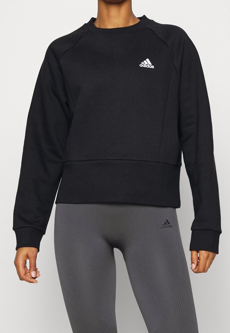 Persona vestida con sudadera negra de manga larga de Adidas y mallas grises de Adidas, de pie contra un fondo claro y liso.