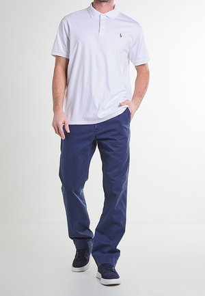 Weißes Poloshirt mit drei Knöpfen und einem kleinen Logo auf der Brust, kombiniert mit dunkelblauen Hose und navyfarbenen Sneakers.