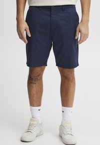 Navy cotton shorts with a flat front, belt loops, and side pockets. Worn with white athletic socks and light-colored sneakers.
