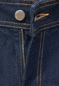 Gros plan d’un jean en denim bleu foncé avec une fermeture éclair métallique partiellement ouverte, des coutures en cuivre et un bouton métallique gravé "Established 1983."