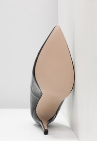 Chaussure à talon haut noire avec une texture façon crocodile, bout pointu et talon fin. La semelle est beige avec un design côtelé, inclinée contre un mur.