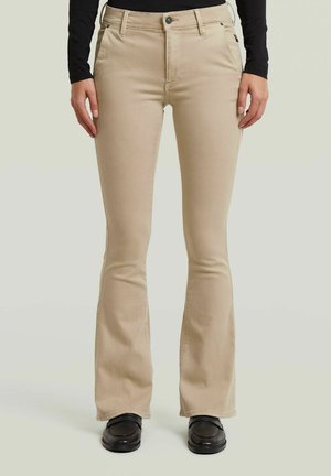Femme portant un pantalon flare beige, un haut noir à manches longues et des chaussures noires, debout, les bras détendus le long du corps.