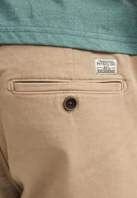 Pantalon en coton beige avec une poche arrière et un détail de bouton foncé. Chemise verte visible en haut, présentant une texture subtile et un ourlet plus court.