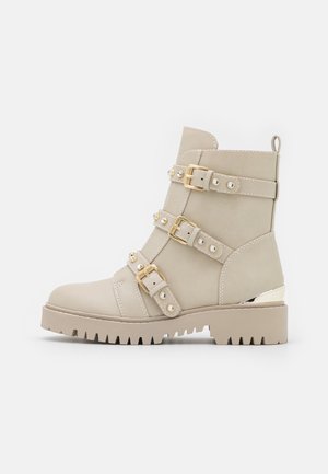 Bottine beige avec trois sangles bouclées dorées et accents de strass, semelle crantée épaisse, et détail métallique sur le talon.