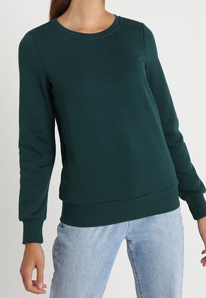 Kobieta w ciemnozielonej bluzie z okrągłym dekoltem, z prążkowanymi mankietami i obszyciem, w połączeniu z jasnoniebieskimi dżinsami na jednolitym tle.