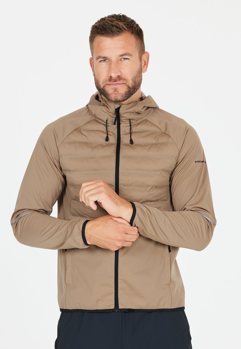 Giacca leggera, di colore beige, imbottita, con chiusura lampo, cappuccio con coulisse, dettagli neri e polsini elastici. Presenta dettagli riflettenti sulle maniche.