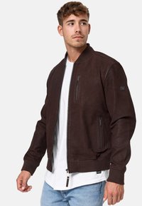 Braune Lederbomberjacke mit gerippten Bündchen und Taillensaum. Verfügt über Seitentaschen und eine Brusttasche mit Reißverschluss. Getragen über einem weißen T-Shirt.