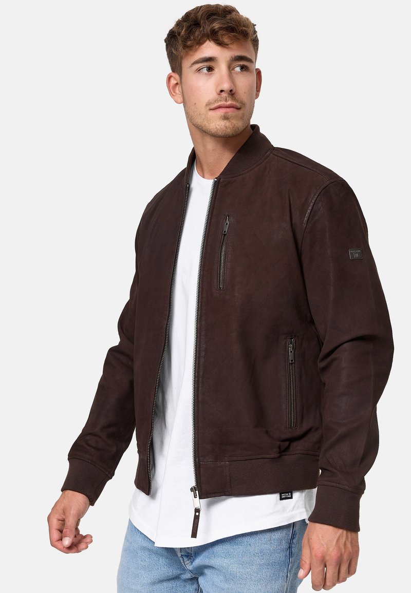Braune Lederbomberjacke mit gerippten Bündchen und Taillensaum. Verf�ügt über Seitentaschen und eine Brusttasche mit Reißverschluss. Getragen über einem weißen T-Shirt.