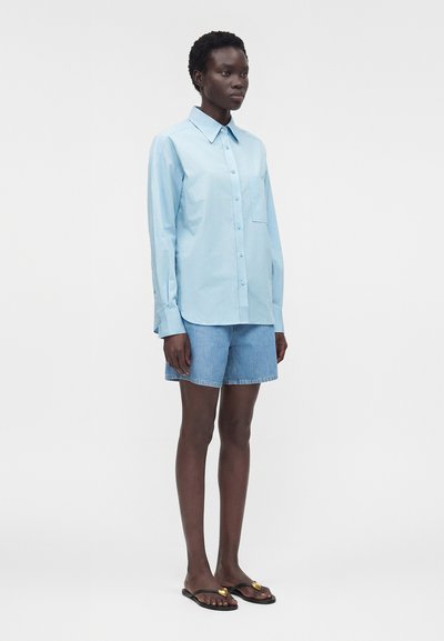 Γυναίκα που στέκεται φορώντας ένα γαλάζιο πουκάμισο με μακριά μανίκια και κουμπιά, τζιν σορτς και μαύρες σαγιονάρες με χρυσές καρδιές ως διακοσμητικά στοιχεία.