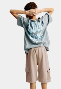 Camiseta gris claro con un gran texto gráfico azul en la parte posterior, combinada con pantalones cortos tipo cargo beige que cuentan con bolsillos laterales y una textura sutil.