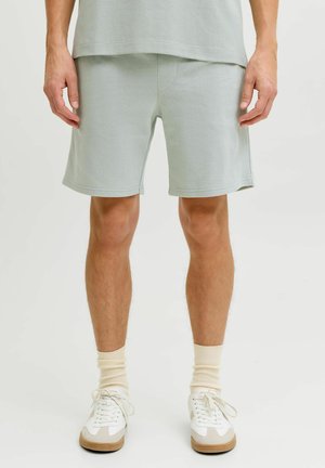 Hombre con pantalones cortos gris claro hasta la rodilla, calcetines blancos a la tripa y zapatillas blancas, de pie sobre un fondo liso.