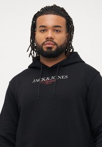 Kübara mees, kellel on dreads ja habe, kannab musta kapuutsiga särk, millel on tekst "JACK & JONES Original Studio" lihtsal valgel taustal.