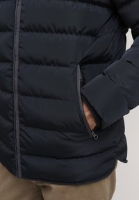 Kék légi puffer dzseki steppelt mintával, cipzáras zsebbel és szürke akcentusokkal a cipzárnál és az ujjaknál. Puha tapintású, párnázott szigeteléssel.