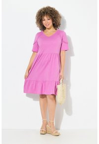Vestido rosa de manga corta con escote en V y bajo de volantes en capas, hecho de tela ligera. Combinado con una bolsa de mimbre y sandalias de esparto.