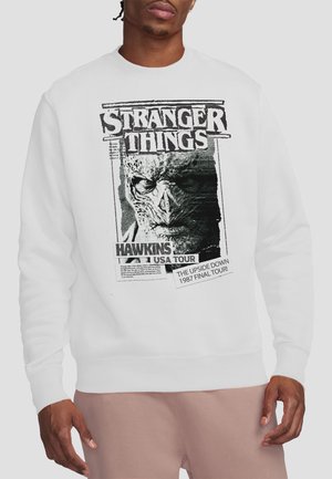 Mann trägt weißen Sweatshirt mit schwarz-weißer Stranger Things-Grafik mit einem Monstergesicht und Text über die Hawkins USA Tour 1987.