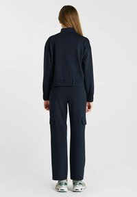 Donker marineblauwe sportset met een jack met ritssluiting en cargobroek met zijzakken. Glad materiaal, relaxte pasvorm en elastische taille.