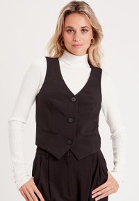Gilet neri con un design aderente, caratterizzati da tre bottoni e due tasche frontali, indossati sopra un top a collo alto a coste bianco. Texture liscia.