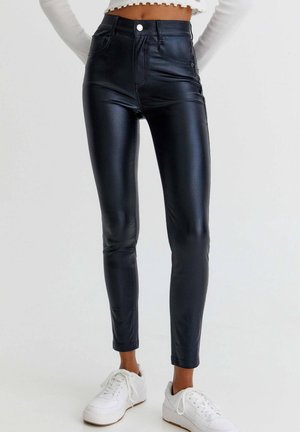 Pantalon noir en similicuir slim porté avec des baskets blanches et un top blanc court à manches longues sur un fond neutre.