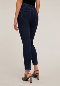 Vaqueros ajustados azul oscuro con una cintura de altura media, que presentan bolsillos traseros y una textura de denim suave. Llevados con sandalias de tacón en piel de serpiente gris.