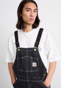 Svarte denimsalopper med justerbare stropper, frontlommer og detaljer i gullhardware. Brukt over en hvit t-skjorte med trykte logoer.