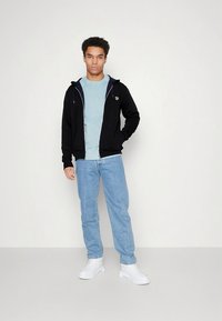 Casaco preto com fecho de correr e logótipo, camisola azul clara por baixo, jeans azul claros e ténis brancos. O conjunto tem um ajuste casual.