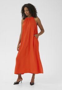 Abito maxi di un vivace arancione, con design a collo halter, realizzato in tessuto morbido. Presenta tasche laterali e una silhouette fluida. Mule nere con tacco.